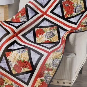 May include: A floral quilt with red, yellow, and purple flowers is draped over a striped armchair. The quilt features a geometric design with black, red, and white borders. The quilt's floral pattern is repeated in square and triangular shapes.