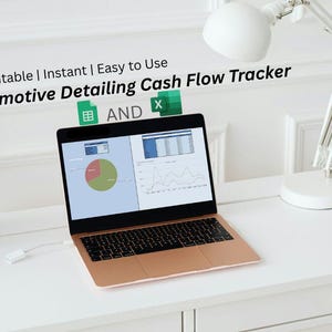 May include: A rose gold laptop displays a cash flow tracker with charts and graphs. The text on the screen reads "Automotive Detailing Cash Flow Tracker". A white desk lamp and a small potted plant are also visible.