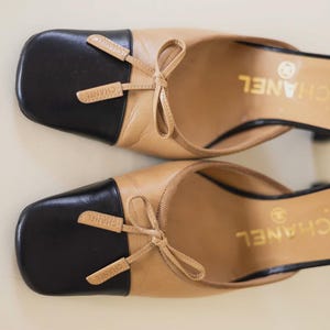Op de afbeelding: Een paar beige en zwarte leren muiltjes met vierkante neuzen. Elke schoen heeft een zwarte neus, een kleine strik en de merknaam "CHANEL" in gouden letters. De hakken zijn zwart.