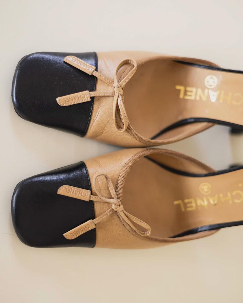 Op de afbeelding: Een paar beige en zwarte leren muiltjes met vierkante neuzen. Elke schoen heeft een zwarte neus, een kleine strik en de merknaam "CHANEL" in gouden letters. De hakken zijn zwart.