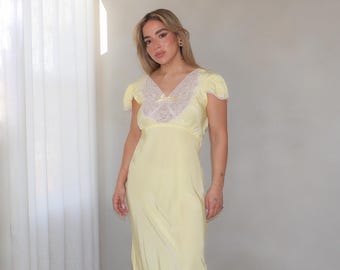 May include: A floor-length pale yellow slip dress featuring short puff sleeves, a V-neckline with white lace trim, and a small central bow. The garment includes a white lace panel at the lower hem and a smooth, flowing fabric construction.