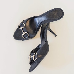 Peut inclure: Une paire de mules à talons noires avec des accessoires argentés. Les chaussures à bout ouvert ont un talon aiguille et une bride sur le dessus du pied. Les chaussures sont en tissu texturé.