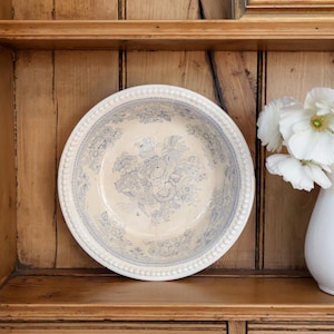 May include: A round ceramic bowl with a cream-colored base and a blue floral transferware pattern. The rim features a raised beaded texture. The bowl is positioned next to a white ceramic pitcher containing white poppy flowers.