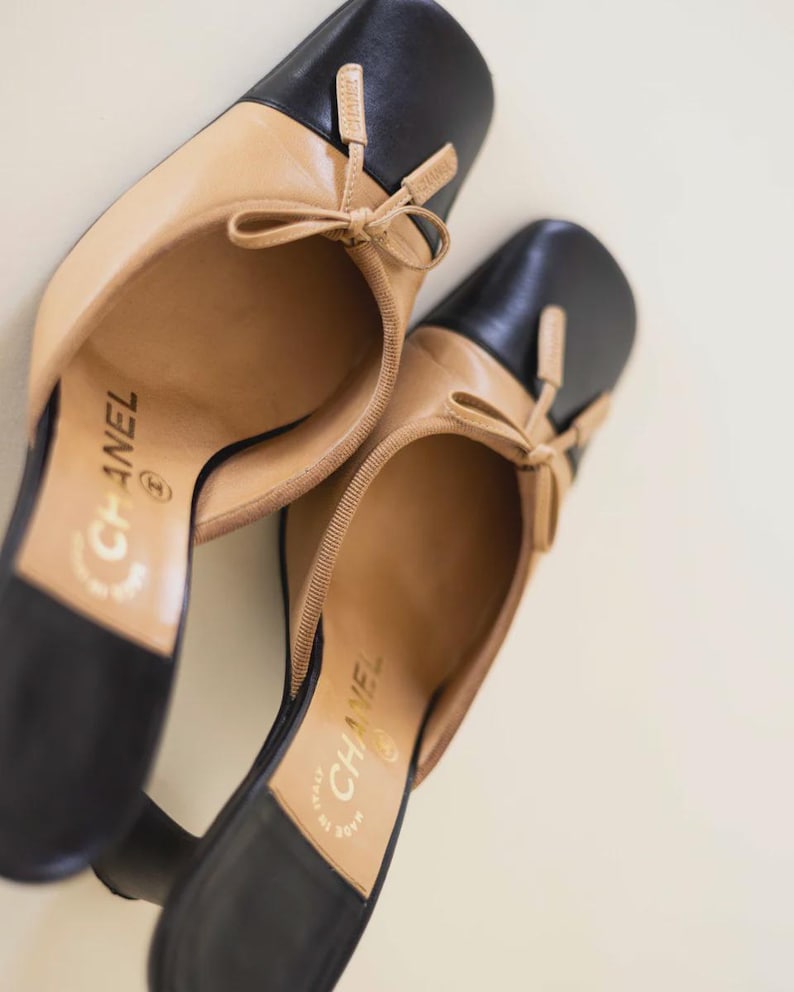 Op de afbeelding: Een paar tweekleurige leren schoenen met hakken. De schoenen hebben een zwarte neus en hiel, met een beige body en een klein strikdetail. De merknaam is zichtbaar op de binnenzool.