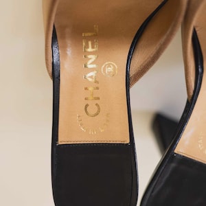 Op de afbeelding: Close-up van een paar beige en zwarte leren sandalen. De binnenzool is bruin met gouden letters met de tekst "CHANEL" en "MADE IN ITALY". De zwarte zool is zichtbaar.