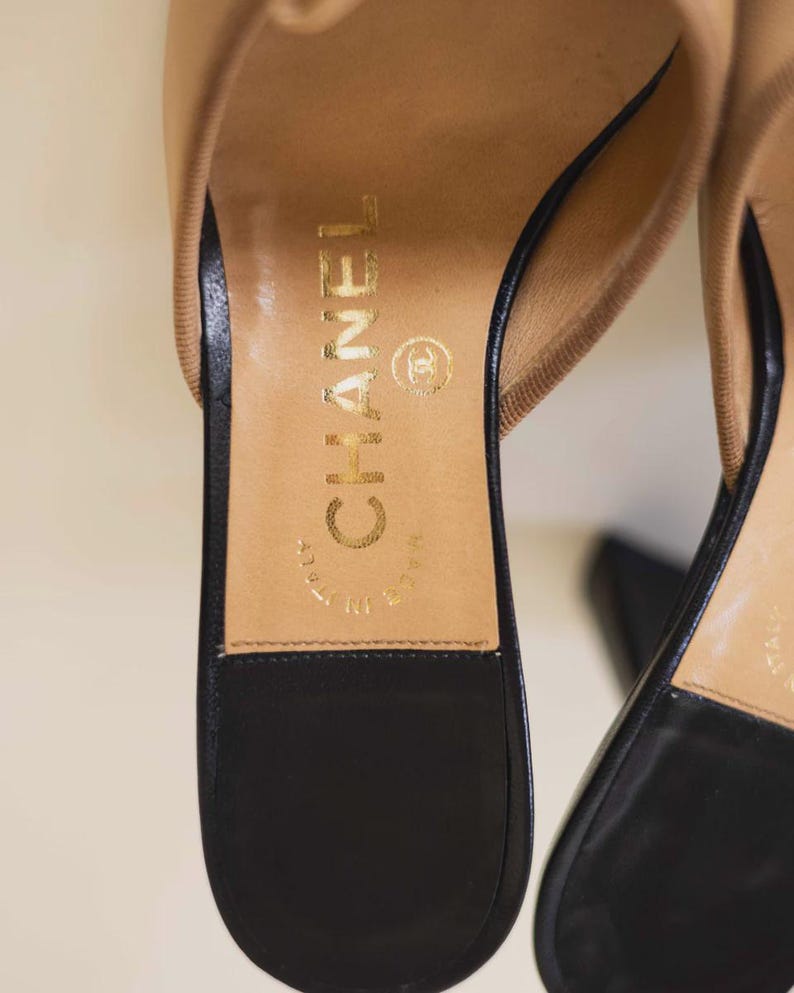 Op de afbeelding: Close-up van een paar beige en zwarte leren sandalen. De binnenzool is bruin met gouden letters met de tekst "CHANEL" en "MADE IN ITALY". De zwarte zool is zichtbaar.