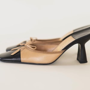 Op de afbeelding: Een paar tweekleurige muiltjes met hakken. De schoenen hebben een zwarte neus en hiel, met een tan bovenwerk en een klein strikdetail. De hakken hebben een uniek, gebogen ontwerp.