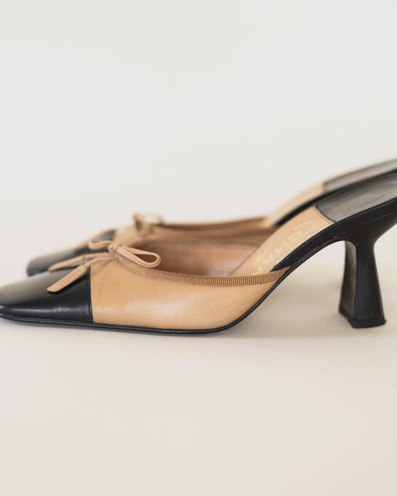 Op de afbeelding: Een paar tweekleurige muiltjes met hakken. De schoenen hebben een zwarte neus en hiel, met een tan bovenwerk en een klein strikdetail. De hakken hebben een uniek, gebogen ontwerp.