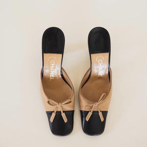Op de afbeelding: Een paar tweekleurige leren muiltjes in beige en zwart. De schoenen hebben een beige bovenwerk met een zwarte neus en hiel. Elke schoen heeft een kleine strik op de wreef. De hakken zijn zwart en de binnenzolen zijn beige.
