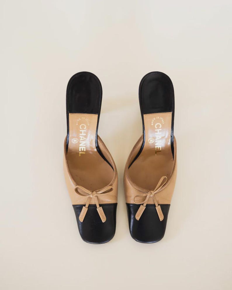Op de afbeelding: Een paar tweekleurige leren muiltjes in beige en zwart. De schoenen hebben een beige bovenwerk met een zwarte neus en hiel. Elke schoen heeft een kleine strik op de wreef. De hakken zijn zwart en de binnenzolen zijn beige.
