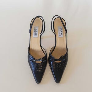 Puede incluir: Un par de zapatos de tacón negros de cuero con punta. Los zapatos tienen un pequeño detalle de lazo y un borde festoneado. La marca "ISAAC" está impresa en la plantilla.