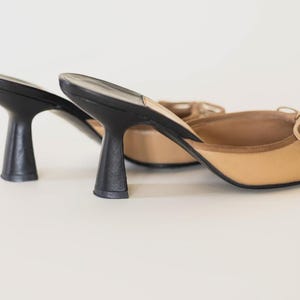 Op de afbeelding: Een paar beige en zwarte muiltjes met hakken. De schoenen hebben een beige bovenwerk met een kleine strik en een zwarte hak. De hakken zijn ongeveer 7,6 cm hoog. De schoenen staan tegen een witte achtergrond.