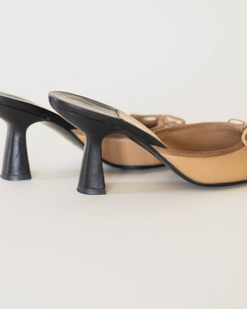 Op de afbeelding: Een paar beige en zwarte muiltjes met hakken. De schoenen hebben een beige bovenwerk met een kleine strik en een zwarte hak. De hakken zijn ongeveer 7,6 cm hoog. De schoenen staan tegen een witte achtergrond.