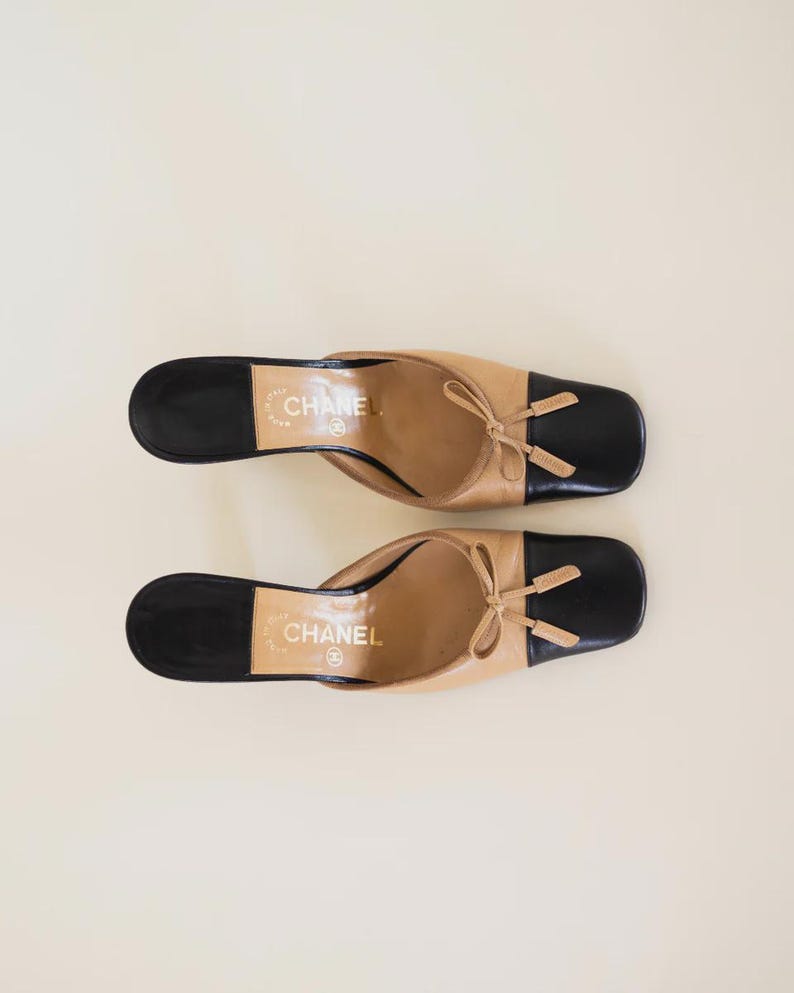 Op de afbeelding: Tweekleurige leren muiltjes in beige en zwart. De schoenen hebben een vierkante neus en een kleine strik. De merknaam "CHANEL" is op de binnenzool gedrukt. Stijlvolle instappers.
