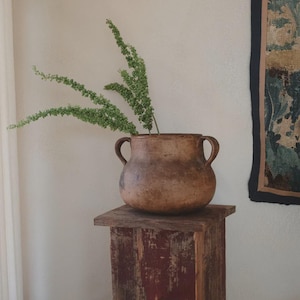 May include: A rustic, brown ceramic vase with two handles, filled with green leafy stems, sits atop a weathered, red-toned wooden pedestal. The vase has a rounded shape and a textured surface. The background includes a wall and a tapestry.