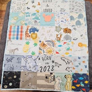 Op de afbeelding: Een patchwork babyquilt met diverse dieren- en patroonvierkanten. De quilt heeft vierkanten met leeuwen, zebra's, olifanten en dinosaurussen. Tekst bevat "LITTLE & LOVED" en "BORN in 2023". De quilt is afgezet met een lichtblauwe stof.