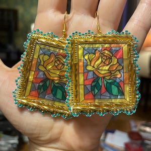 May include: Square earrings with a stained glass design featuring a yellow rose with green leaves. The earrings have a gold-coloured frame and are trimmed with small turquoise beads. The background of the design includes red, orange, and yellow.