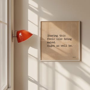Peut inclure: Une affiche encadrée avec le texte "Sharing this feels like being naked. Might as well be." L'affiche est sur un fond beige avec un cadre blanc. Une lampe murale orange est fixée au mur.