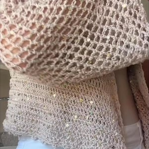 May include: A beige crochet cardigan featuring an open-knit pattern and scattered small, reflective sequins throughout the fabric. The garment has long sleeves and a textured, lightweight construction with a scalloped hemline along the bottom edge.
