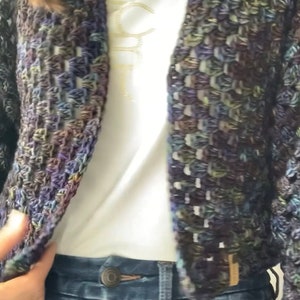 Puede incluir: Primer plano de una mujer con un cárdigan de crochet azul oscuro y verde con una camiseta blanca debajo y unos vaqueros azules.