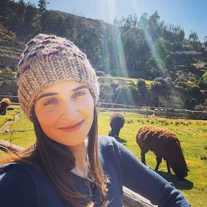 May include: A woman wearing a beige knitted hat with purple accents and a blue sweater, standing in a field with a brown and black sheep in the background.
