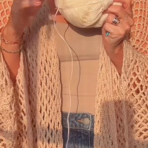 May include: A cream-colored, open-weave crochet cardigan with wide sleeves. A ball of cream-colored yarn is held in the hands, with a crochet hook visible. The cardigan is paired with a neutral-colored top and blue jeans.
