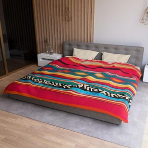 May include: A bedspread featuring a vibrant horizontal striped pattern in shades of red, teal, yellow, and black with geometric accents. The textile covers a gray upholstered bed frame with two cream-colored rectangular pillows placed at the head.