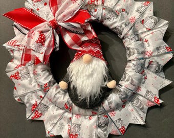 May include: A white and red Christmas wreath with a gnome in the center. The wreath is made of ribbon with a pattern of gnomes and snowflakes. The gnome is wearing a red and white hat and a black and white outfit. A red and white checkered bow is on top of the wreath.