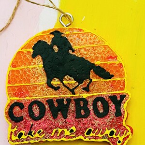May include: A round, glittery, orange and pink ornament with a black silhouette of a cowboy riding a horse. The ornament has the text "COWBOY take me away" written on it.