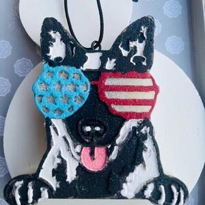 May include: A black and white dog ornament with blue star-shaped sunglasses on the left and red and white striped sunglasses on the right. The dog has a pink tongue sticking out and a black string for hanging.