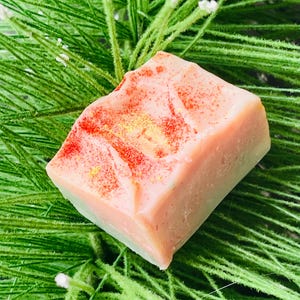 Op de afbeelding: Een vierkant stuk zeep met een lichtroze bovenkant en een lichtgroene onderkant. De bovenkant heeft rode en gouden glitteraccenten. De zeep ligt op een bedje van groen, naaldachtig blad met kleine witte bloemen.