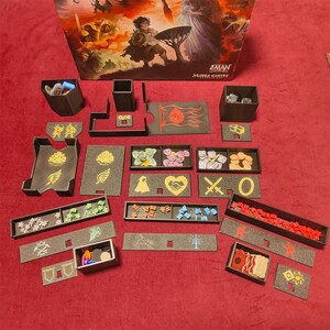 Könnte beinhalten: Ein Brettspiel-Set mit verschiedenen Komponenten, darunter schwarze Tabletts, Spielsteine und eine Box mit Kunstwerken. Die Spielsteine sind in verschiedenen Farben gehalten, und die Tabletts haben goldene und weiße Designs. Die Box zeigt eine Fantasy-Landschaft.