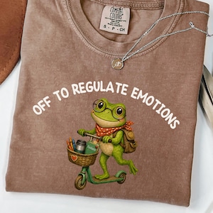 Op de afbeelding: Een bruin t-shirt met een afbeelding van een groene kikker met een bril en een rode sjaal met stippen, rijdend op een step met een mandje met kleurpotloden en een boek. De tekst "OFF TO REGULATE EMOTIONS" staat in het wit boven de kikker.