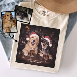 May include: A cream-colored t-shirt featuring a graphic print of two dogs wearing Santa hats, sitting at a bar with glasses of beer. The text 'tis the season' is printed below the dogs. Two smaller inset photos of the dogs are shown above.