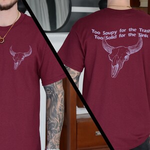 May include: A maroon t-shirt with a white bull skull graphic on the front and the text "Too Soupy for the Trash Too Solid for the Sink" on the back.
