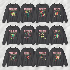 May include: Twelve gray sweatshirts with colorful zodiac signs and names. The signs are Capricorn, Aquarius, Pisces, Aries, Taurus, Gemini, Cancer, Leo, Virgo, Libra, Scorpio, and Sagittarius.