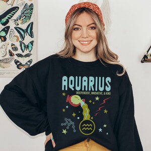K&ouml;nnte beinhalten: Schwarzer Pullover mit einem grafischen Design des Sternzeichens Wassermann. Das Design zeigt einen blauen und gr&uuml;nen Wassermann mit Sternen und dem Text "Wassermann, unabh&auml;ngig, innovativ und freundlich".