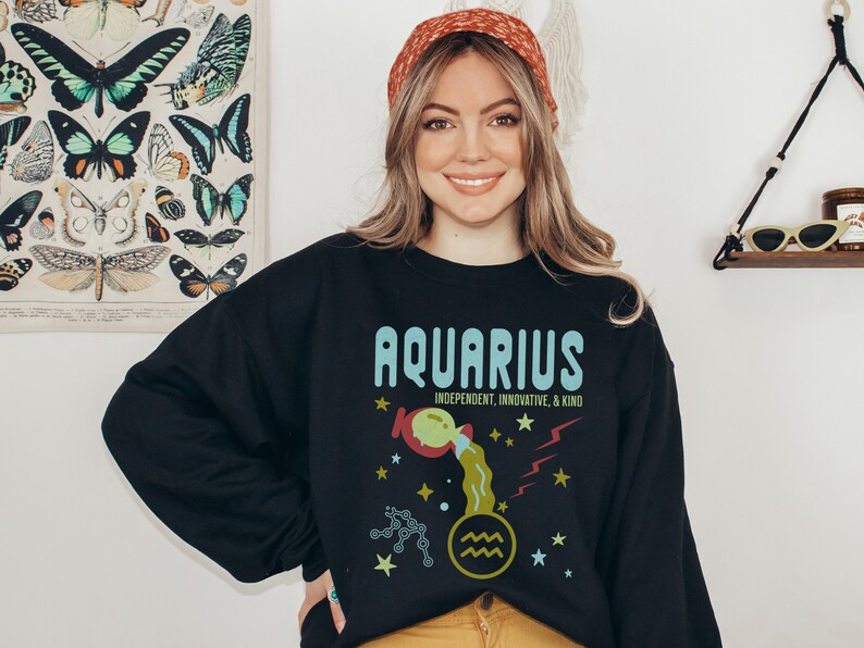 K&ouml;nnte beinhalten: Schwarzer Pullover mit einem grafischen Design des Sternzeichens Wassermann. Das Design zeigt einen blauen und gr&uuml;nen Wassermann mit Sternen und dem Text "Wassermann, unabh&auml;ngig, innovativ und freundlich".