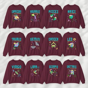 May include: Twelve maroon sweatshirts with a different zodiac sign on each. The signs are Capricorn, Aquarius, Pisces, Aries, Taurus, Gemini, Cancer, Leo, Virgo, Libra, Scorpio, and Sagittarius. Each sign is depicted with a colorful illustration and the name of the sign in blue text.