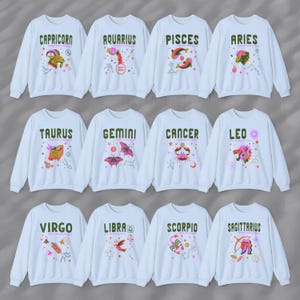 May include: Twelve white sweatshirts with colorful zodiac signs and symbols printed on the front. The signs are: Capricorn, Aquarius, Pisces, Aries, Taurus, Gemini, Cancer, Leo, Virgo, Libra, Scorpio, and Sagittarius.