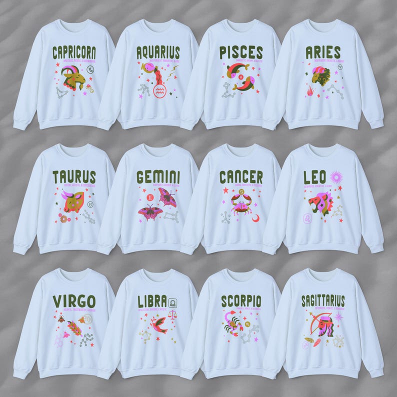 May include: Twelve white sweatshirts with colorful zodiac signs and symbols printed on the front. The signs are: Capricorn, Aquarius, Pisces, Aries, Taurus, Gemini, Cancer, Leo, Virgo, Libra, Scorpio, and Sagittarius.