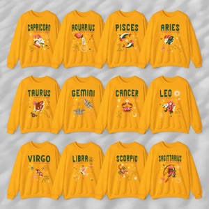May include: Twelve yellow sweatshirts with zodiac signs printed on the front. Each sweatshirt features a different zodiac sign with a unique illustration and text. The signs include Capricorn, Aquarius, Pisces, Aries, Taurus, Gemini, Cancer, Leo, Virgo, Libra, Scorpio, and Sagittarius.