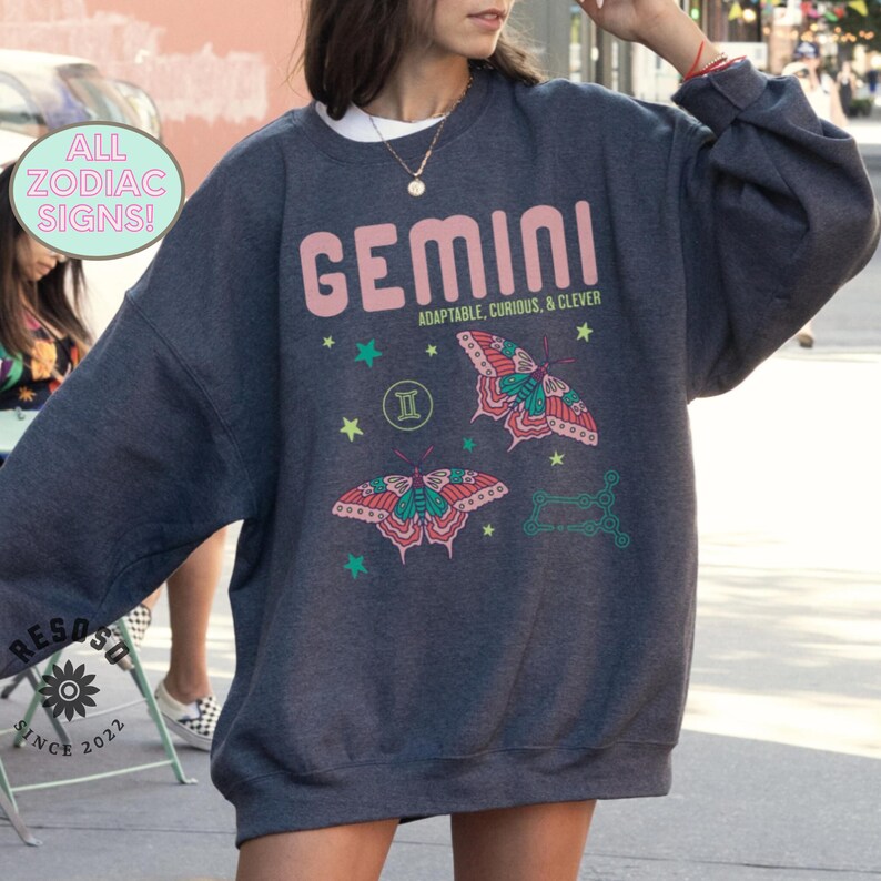 May include: A dark gray sweatshirt with a pink graphic of the Gemini zodiac sign, two butterflies, and stars. The text "GEMINI" is in pink with the words "ADAPTABLE, CURIOUS, & CLEVER" in smaller font below.