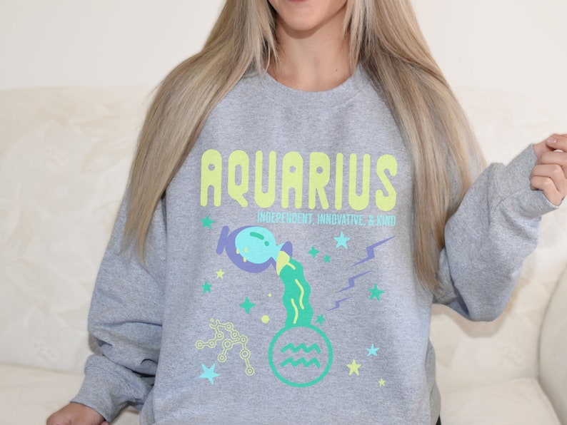 K&ouml;nnte beinhalten: Grauer Pullover mit einer bunten Grafik des Sternzeichens Wassermann. Die Grafik enth&auml;lt das Wort "Wassermann" in Gelb, eine blaue und gr&uuml;ne Wasser gie&szlig;ende Figur und die Worte "Unabh&auml;ngig, Innovativ & Freundlich".