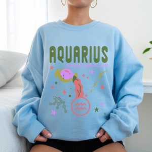 K&ouml;nnte beinhalten: Hellblauer Pullover mit einem grafischen Design des Sternzeichens Wassermann. Das Design zeigt einen rosa und orangefarbenen Wasserkrug mit Sternen und Blitzen. Der Text "Aquarius" ist in gr&uuml;nen Buchstaben geschrieben.