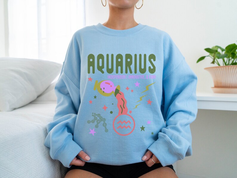 K&ouml;nnte beinhalten: Hellblauer Pullover mit einem grafischen Design des Sternzeichens Wassermann. Das Design zeigt einen rosa und orangefarbenen Wasserkrug mit Sternen und Blitzen. Der Text "Aquarius" ist in gr&uuml;nen Buchstaben geschrieben.