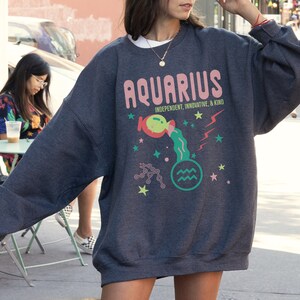 K&ouml;nnte beinhalten: Ein dunkelgrauer Pullover mit einer bunten Grafik des Sternzeichens Wassermann. Die Grafik enth&auml;lt das Wort "Wassermann" in rosa Buchstaben, ein Cartoon-Bild eines Wasserkruges, der Wasser gie&szlig;t, und die Worte "Unabh&auml;ngig, Innovativ und Freundlich".
