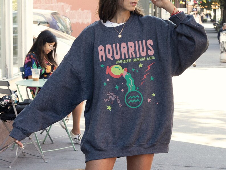 K&ouml;nnte beinhalten: Ein dunkelgrauer Pullover mit einer bunten Grafik des Sternzeichens Wassermann. Die Grafik enth&auml;lt das Wort "Wassermann" in rosa Buchstaben, ein Cartoon-Bild eines Wasserkruges, der Wasser gie&szlig;t, und die Worte "Unabh&auml;ngig, Innovativ und Freundlich".