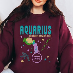 K&ouml;nnte beinhalten: Ein weinroter Pullover mit einem grafischen Design, das das Sternzeichen Wassermann zeigt. Das Design beinhaltet einen t&uuml;rkisblauen und gr&uuml;nen Text, der "Aquarius" sagt, mit den Worten "Unabh&auml;ngig, Innovativ & Freundlich" darunter. Die Grafik enth&auml;lt auch einen lila und gr&uuml;nen Wasserkrug, der Wasser gie&szlig;t, ein lila Wassermann-Symbol und goldene und blaue Sterne.