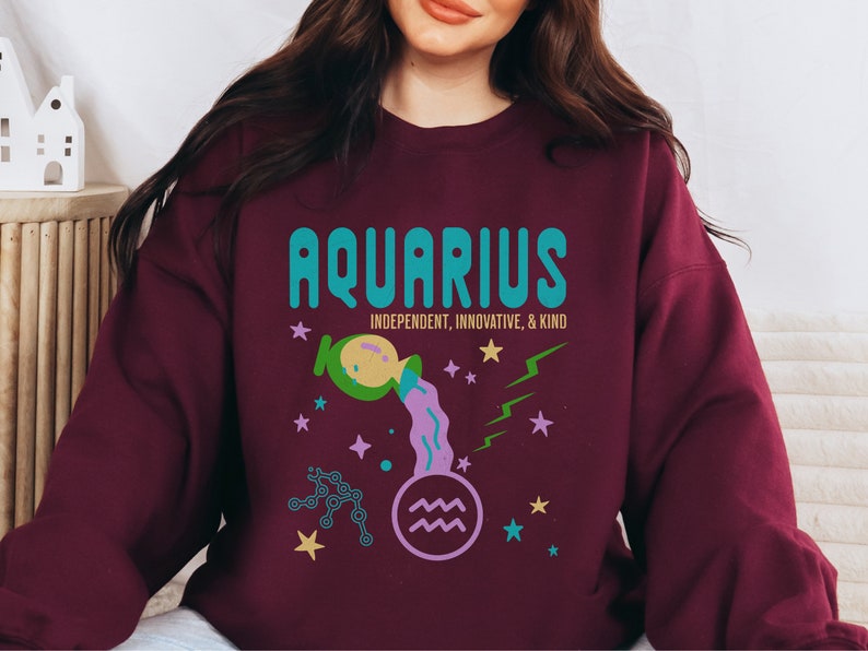 K&ouml;nnte beinhalten: Ein weinroter Pullover mit einem grafischen Design, das das Sternzeichen Wassermann zeigt. Das Design beinhaltet einen t&uuml;rkisblauen und gr&uuml;nen Text, der "Aquarius" sagt, mit den Worten "Unabh&auml;ngig, Innovativ & Freundlich" darunter. Die Grafik enth&auml;lt auch einen lila und gr&uuml;nen Wasserkrug, der Wasser gie&szlig;t, ein lila Wassermann-Symbol und goldene und blaue Sterne.